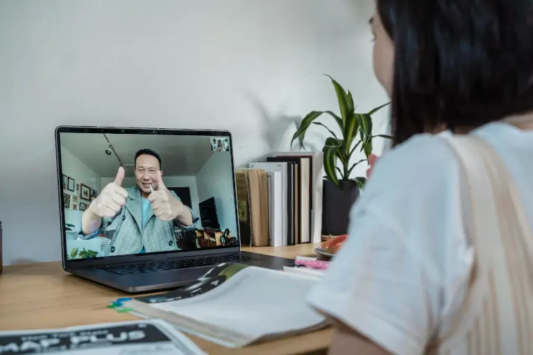 A student sits at a desk with study materials, engaging in an online tutoring session on a laptop where the tutor gives a double thumbs-up gesture.